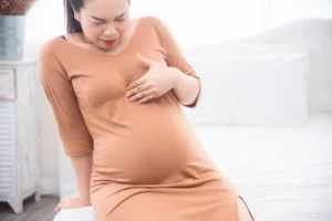 Asian pregnant woman sitting on bed suffering from heartburn in third trimester. 