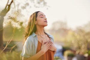 Young Latin woman with hand on chest breathing in fresh air in a beautiful garden during sunset. Healthy Mexican girl enjoying nature while meditating during morning exercise routine with closed eyes. 