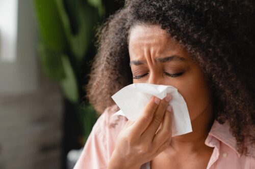 Sneezing coughing ill young Arican woman using paper napkin, having runny nose, blowing her nose. Coronavirus, infectious disease, flu, cold.