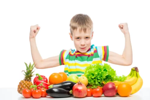 small strongman shows biceps at the table with a pile of fresh vegetables and fruits isolated in the studio.