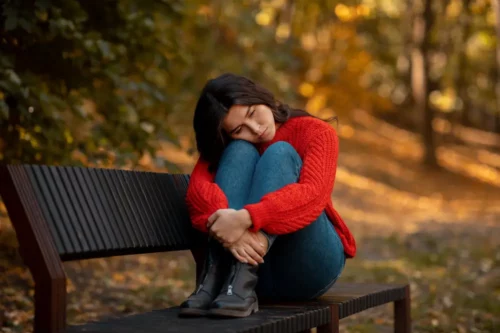 Depressed young woman hugging her knees while sitting on bench at yellow autumn park. Millennial lady feeling stressed or unhappy, suffering from loneliness or seasonal affective disorder, outdoors.
